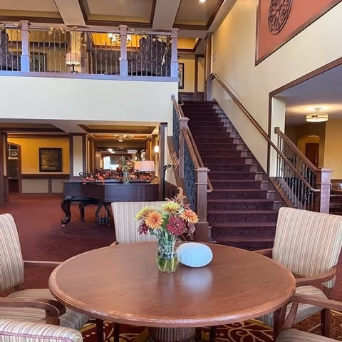 Lobby seating area with a round table and vase of flowers, striped chairs, a grand piano, and a carpeted staircase leading to an upper level.