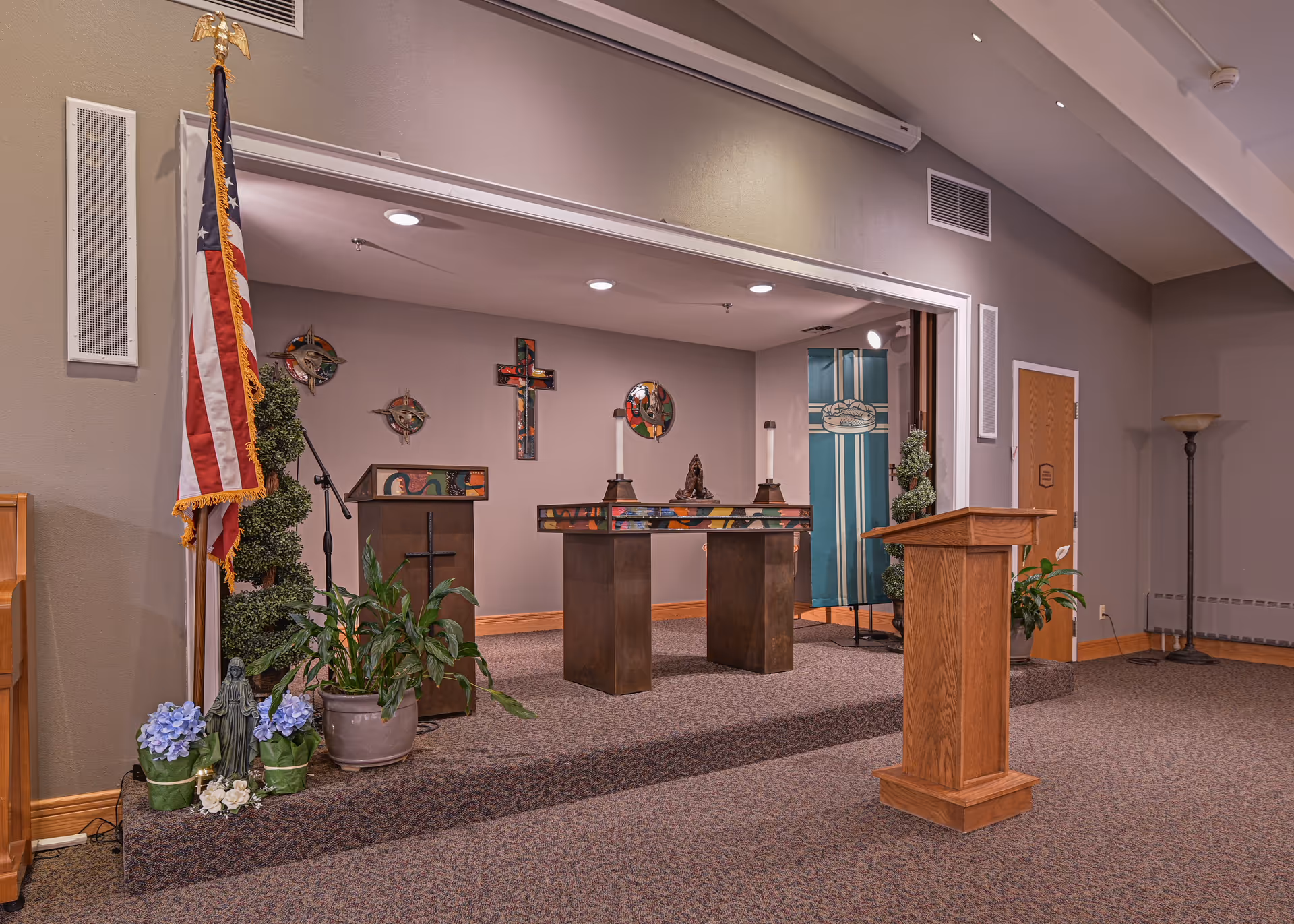 Interior view of a chapel or worship area with a wooden podium, altar table with candles, religious crosses and decorations on the wall, an American flag, potted plants, and a microphone stand.