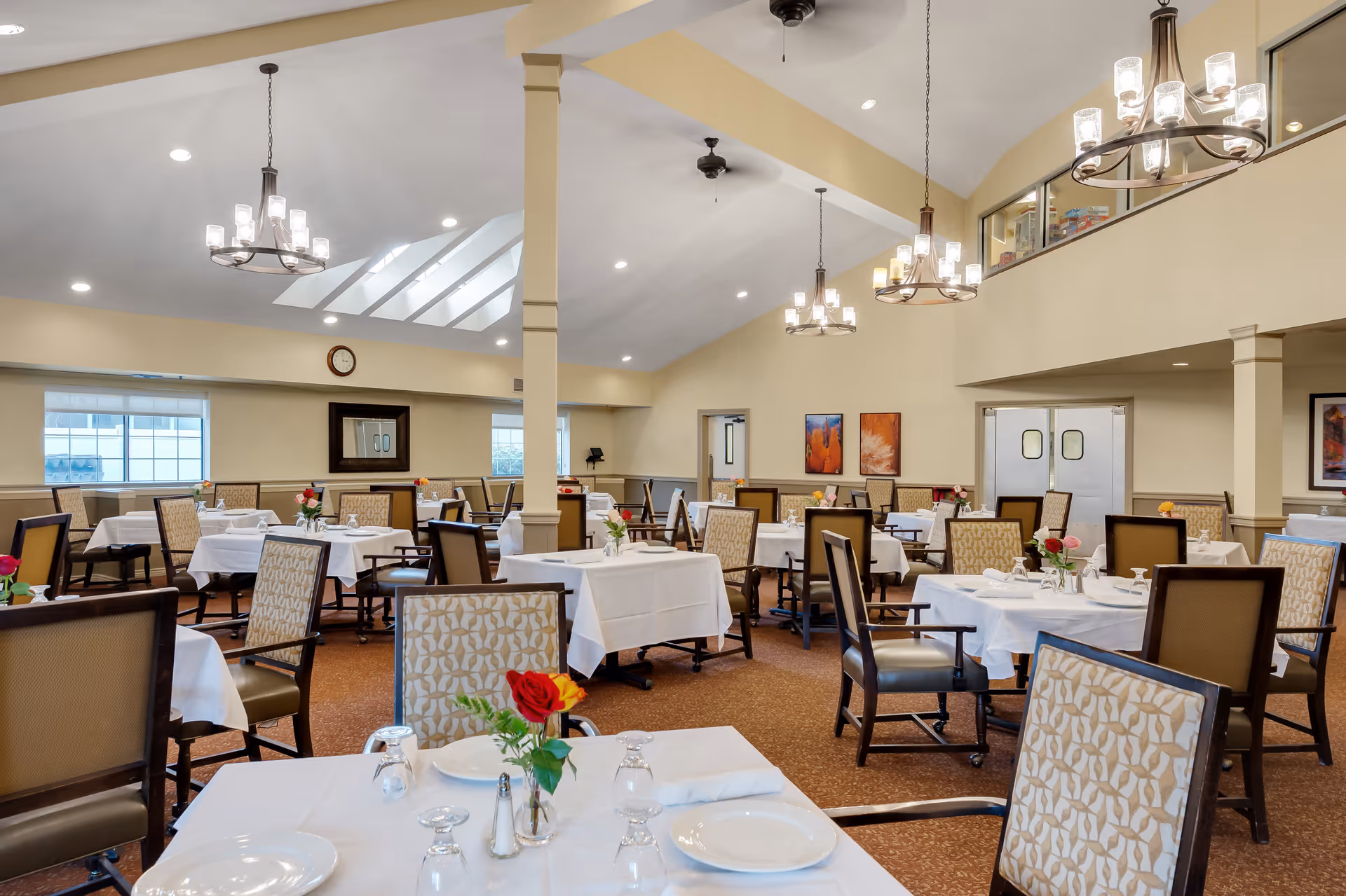 A spacious dining room with multiple tables covered in white tablecloths, each set with plates, glasses, napkins, and small floral centerpieces. The room features high ceilings with chandeliers and ceiling fans, beige walls, and large windows allowing natural light to enter. There are patterned upholstered chairs around the tables and framed artwork on the walls.