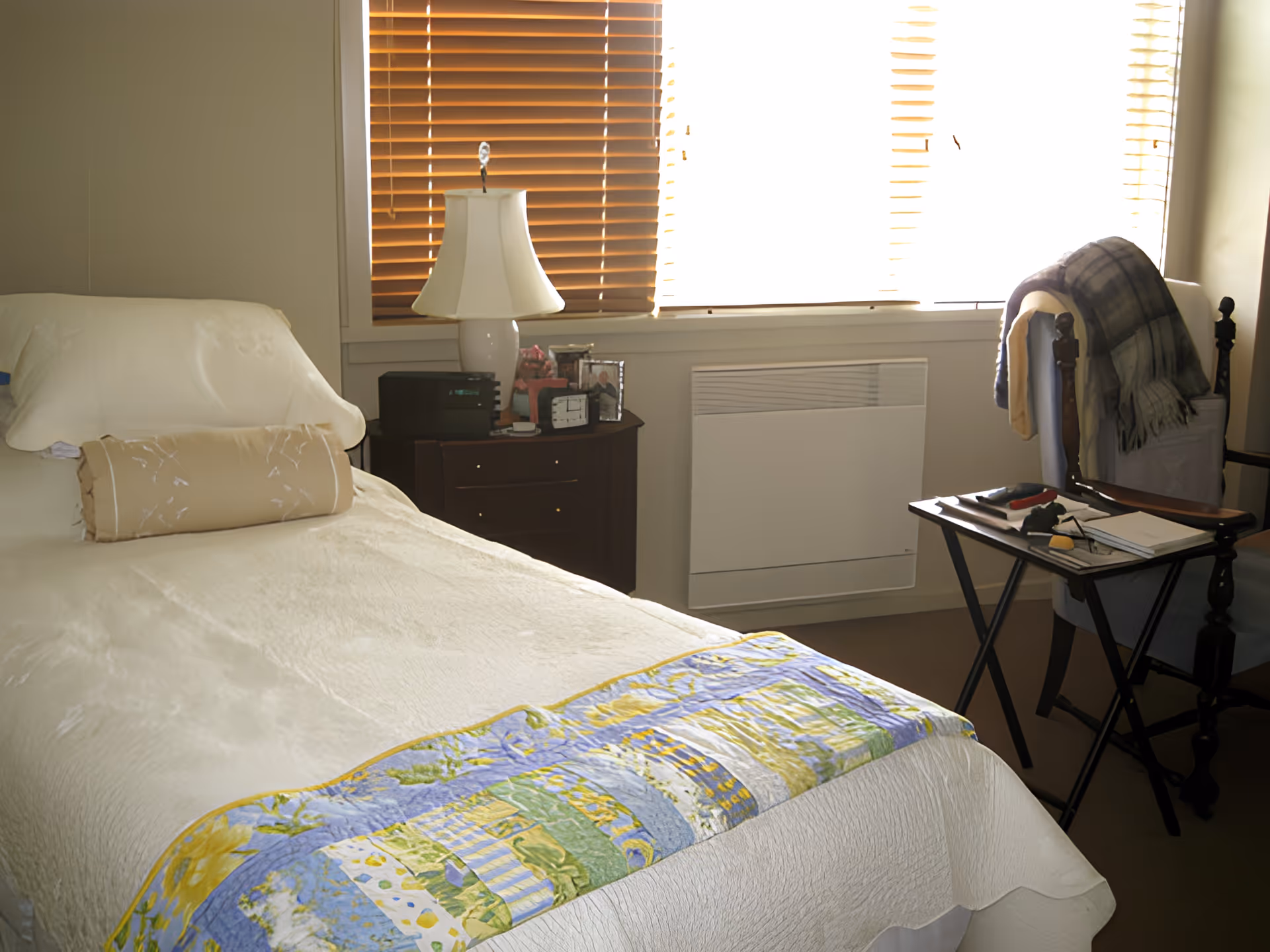A cozy bedroom with a single bed covered in a cream-colored bedspread and a decorative pillow. A colorful quilt is folded at the foot of the bed. Next to the bed is a dark wooden nightstand with a white lamp, a clock, and some framed photos. A window with wooden blinds lets in bright light. There is a chair with a plaid blanket draped over it and a small folding table holding various items including a notebook and glasses.