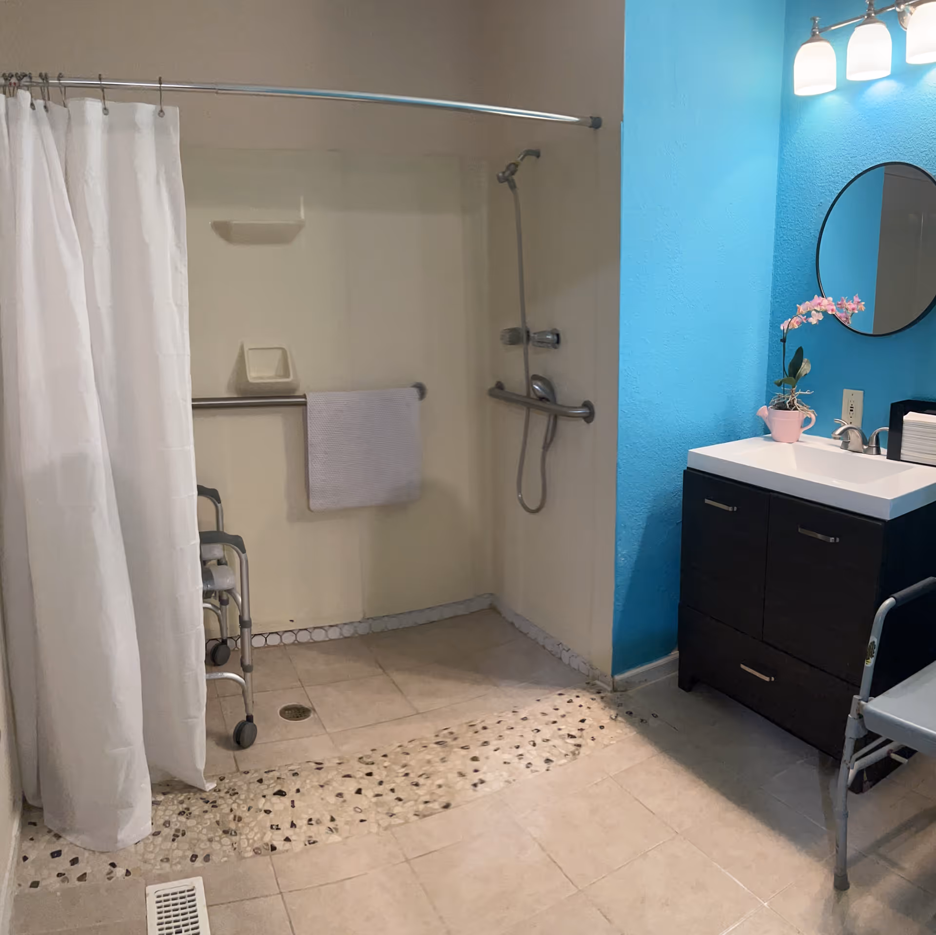 Accessible bathroom with a walk-in shower featuring a handheld showerhead, grab bars, and a white shower curtain. A towel hangs on a grab bar inside the shower. There is a dark wood vanity with a white sink, a round mirror above it, and a small pink flower pot with a blooming orchid on the counter. The wall behind the vanity is painted bright blue. A walker is partially visible near the shower.