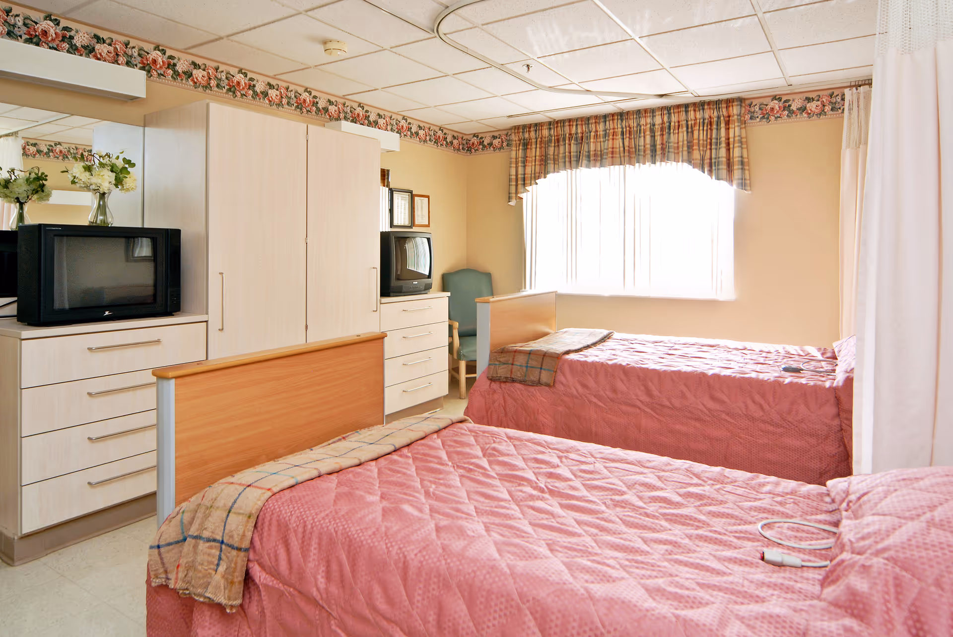 Two twin beds with pink bedspreads in a well-lit senior living bedroom with dressers, TVs, and a window.