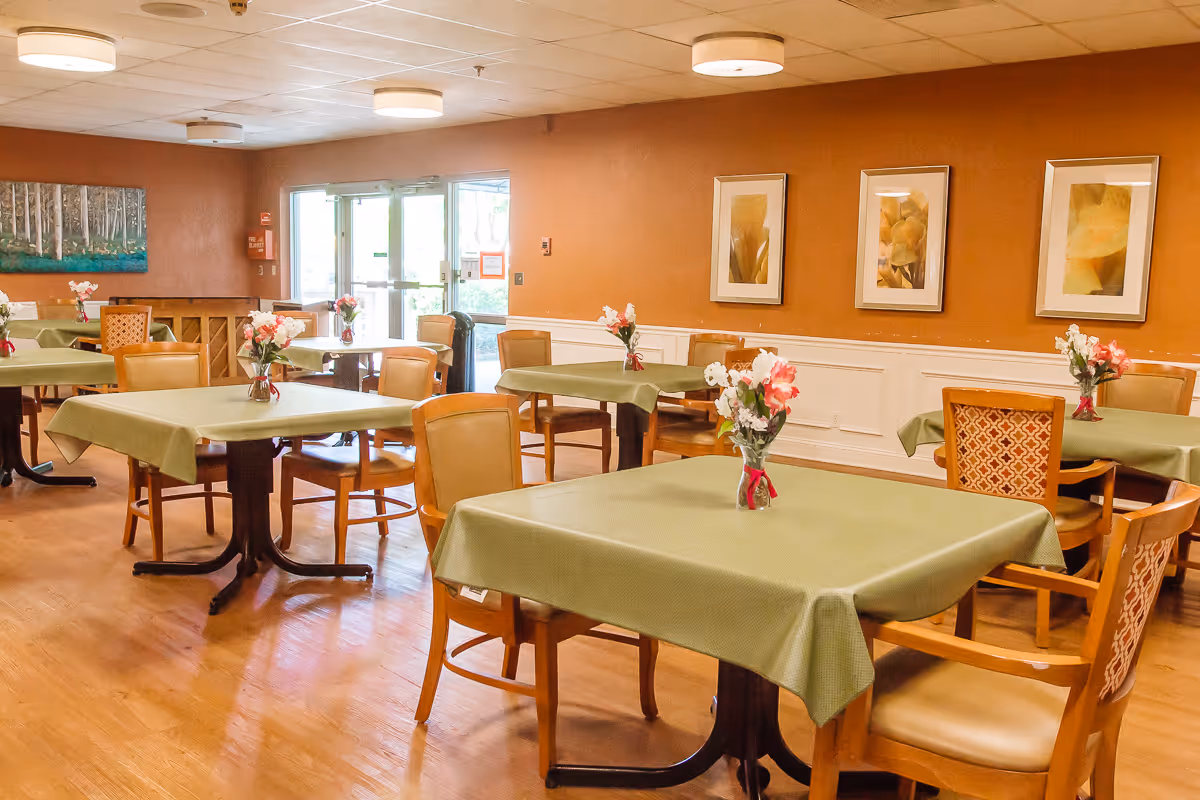 A dining room with several square tables covered with green tablecloths, each table decorated with a small vase of flowers. The room has wooden chairs, hardwood floors, warm-colored walls with framed floral artwork, and a glass door leading outside.