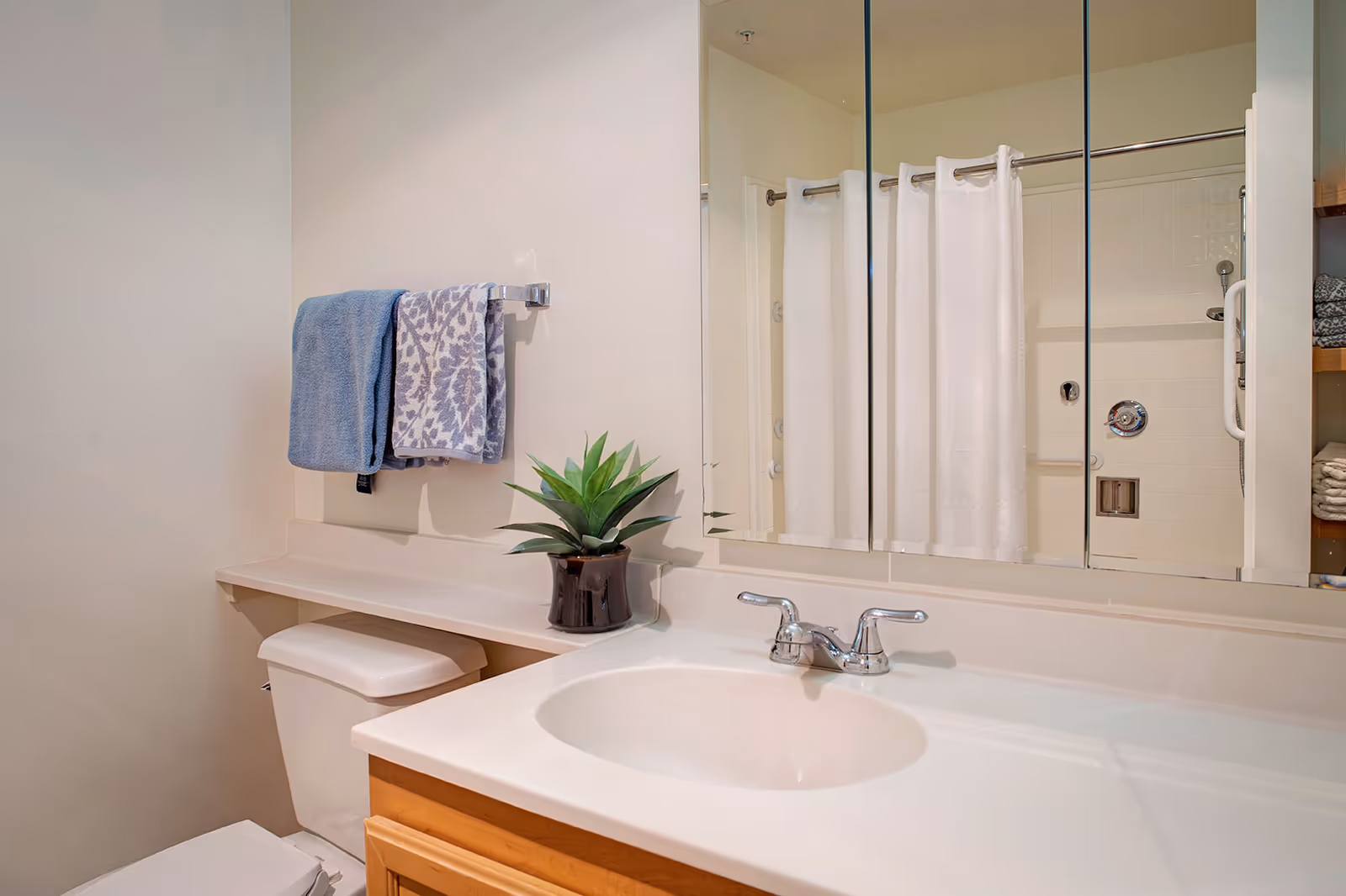 A clean bathroom featuring a white sink with a silver faucet, a toilet, a small potted plant on the counter, two towels hanging on a towel rack, and a large mirror reflecting a shower area with a white curtain.