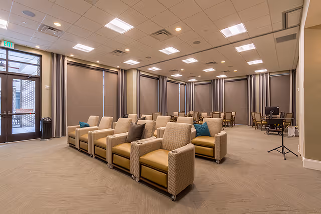 Spacious lounge with rows of upholstered recliners, tables and chairs in the back, and large windows with shades.