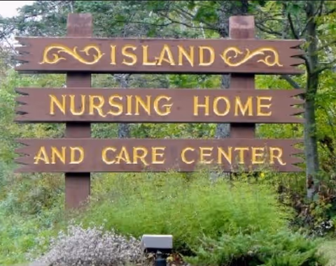 Wooden roadside sign that reads "ISLAND NURSING HOME AND CARE CENTER" set among green foliage.