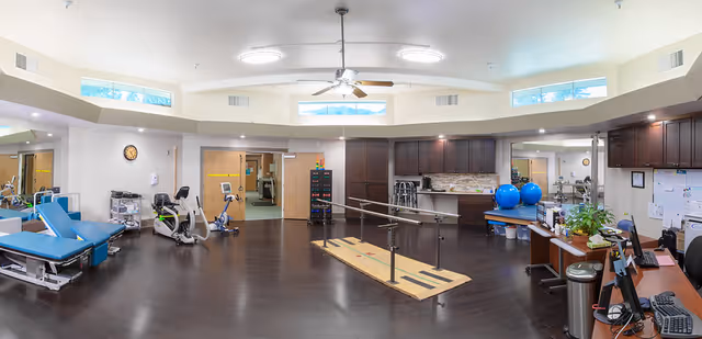 A spacious rehabilitation or physical therapy room with dark wooden floors and white walls. The room contains exercise equipment including stationary bikes, parallel bars for walking practice, therapy tables, large blue exercise balls, and a desk area with a computer and office supplies. There are several windows near the ceiling allowing natural light in, and ceiling fans for ventilation.
