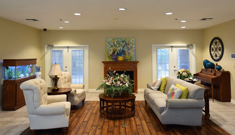A bright common living room with upholstered sofa and armchairs arranged around a wooden coffee table topped with a floral arrangement, an aquarium, piano, and double glass doors.