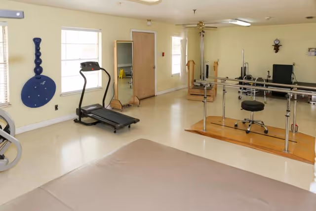 Bright rehabilitation room with a treadmill, parallel bars, therapy equipment, and a mirror near windows.