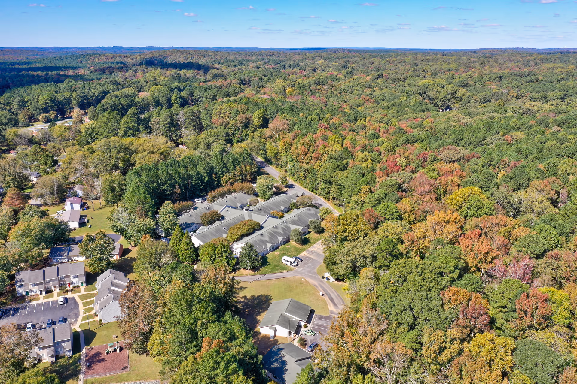 Aerial view of Cambridge Hills Assisted Living facility surrounded by dense forest with trees showing early fall colors under a clear blue sky.