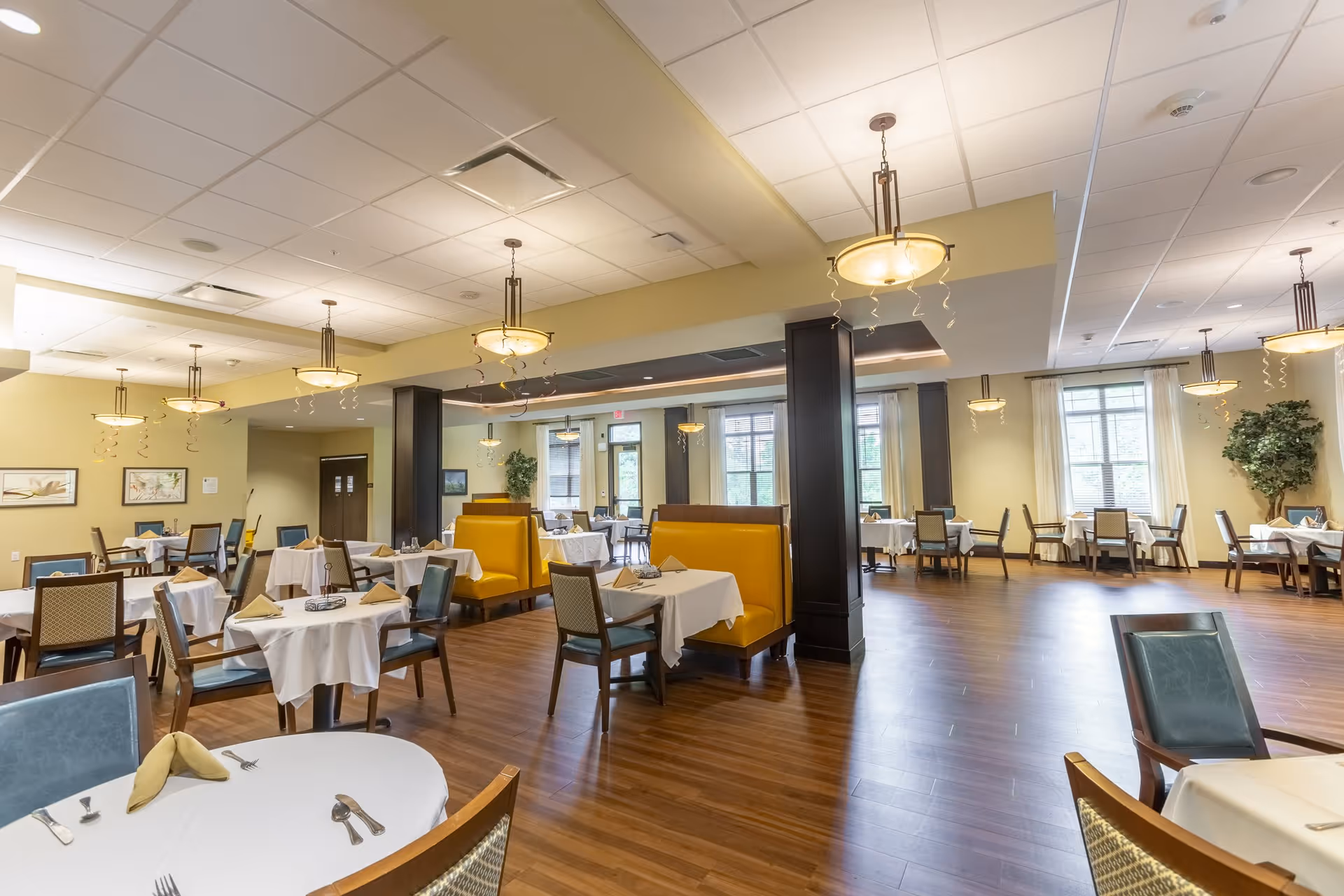 A spacious dining room with multiple tables covered in white tablecloths, set with silverware and napkins. The room features wooden flooring, yellow booth seating, several chairs, large windows with white curtains, and modern hanging light fixtures.