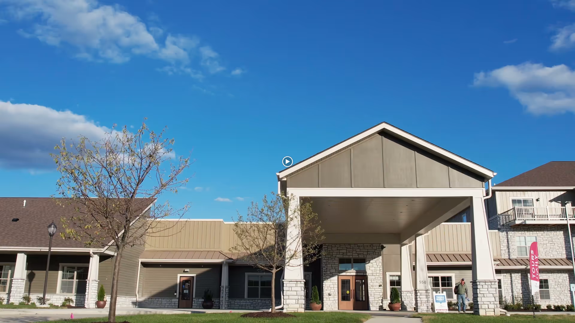Front entrance of a senior living building with a covered porte-cochère, stone columns, small trees, and a clear blue sky.