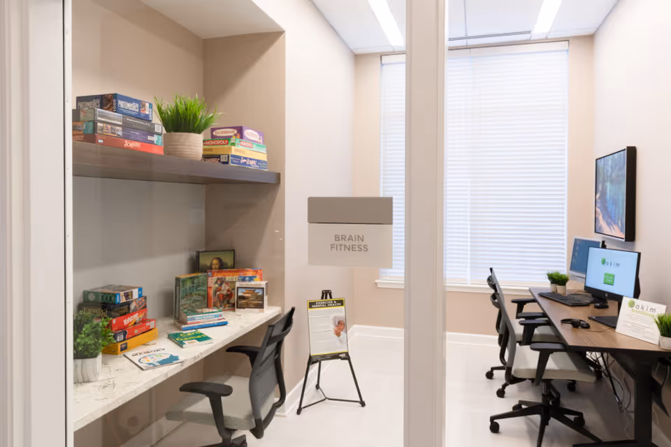 A bright room labeled 'Brain Fitness' with shelves holding board games and puzzles on the left side, and a desk with computers and chairs on the right side. The room has large windows with blinds, light-colored walls, and a clean, organized appearance.