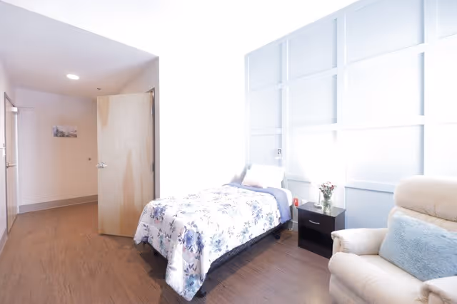 Bright private bedroom with a single floral bed, nightstand, recliner chair, and an open door.