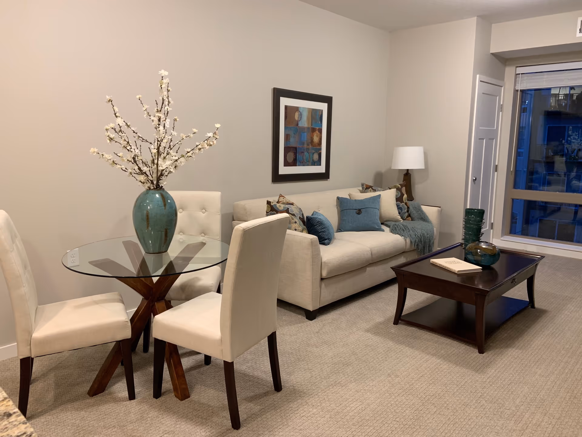 A cozy living room area with a beige sofa adorned with blue and patterned cushions, a wooden coffee table with decorative items and a book, a floor lamp, and a glass-top round dining table with four cream-colored upholstered chairs. A large window with a view of a building outside is visible in the background.
