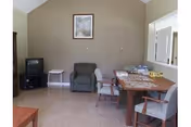 A common room with a wooden table and four chairs, a green armchair, a small white side table, and a TV on a stand in the corner. There is a framed picture on the beige wall above the armchair.