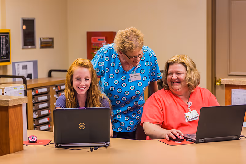 Three women in a nursing facility office setting, two seated at a counter working on laptops and one standing behind them smiling. The woman on the left has red hair and is using a Dell laptop, the woman in the middle is wearing a blue patterned scrub top, and the woman on the right is wearing a red scrub top. They appear to be engaged in a friendly conversation.