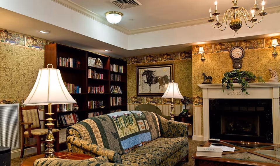A cozy common room with a patterned sofa, table lamps, bookshelves, framed art, and a fireplace under a chandelier.