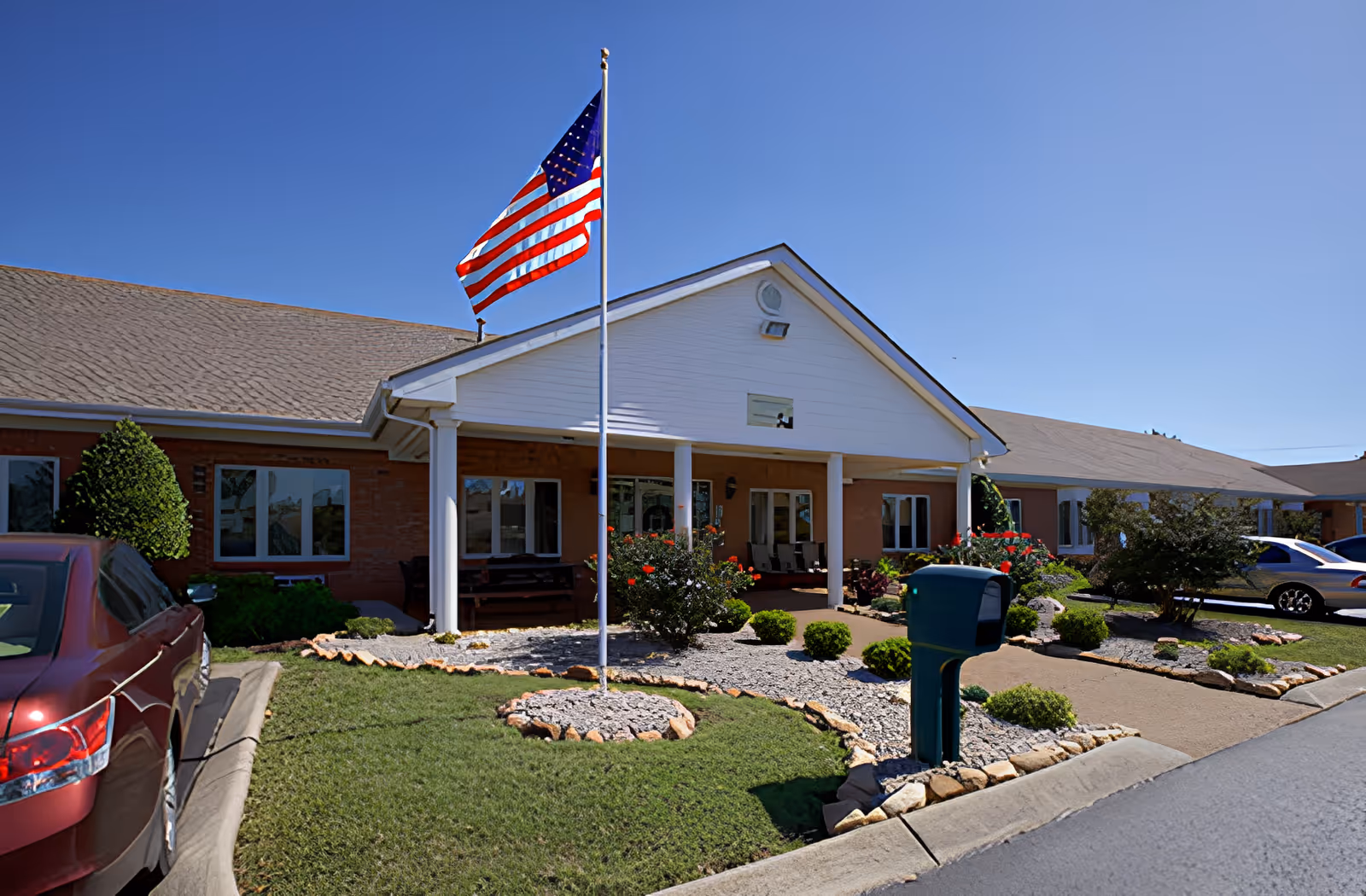 Exterior view of a single-story brick building with a covered entrance supported by white columns. An American flag is flying on a flagpole in front of the building, surrounded by landscaped bushes and flowers. Several cars are parked along the driveway and parking area.