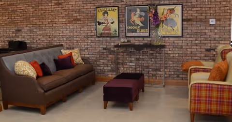 A cozy living room with a brown sofa adorned with colorful pillows, a purple ottoman, and a plaid armchair with orange cushions. The background features a brick wall decorated with three vintage-style framed posters and a small table with a vase of flowers.