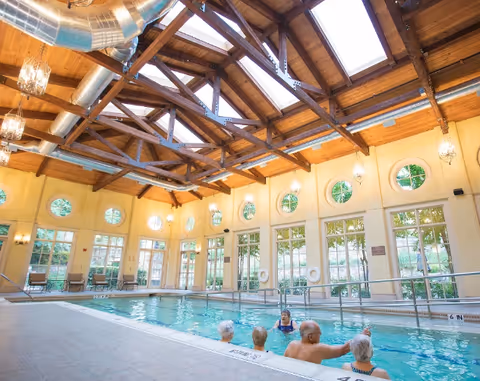 Indoor swimming pool with several elderly people enjoying the water. The pool area features large windows with circular and rectangular panes, wooden ceiling beams, skylights, and hanging light fixtures. There are chairs along the walls and greenery visible outside the windows.