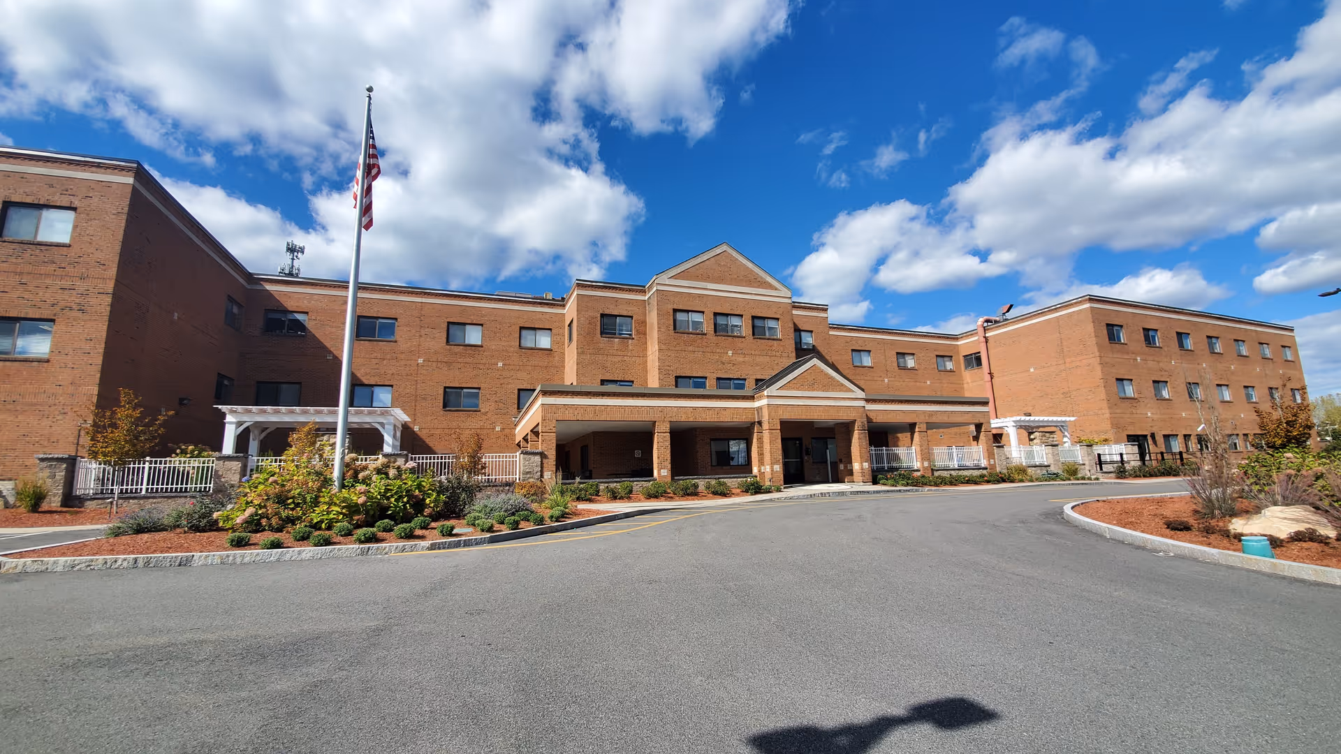 Front exterior view of a large brick senior living facility building with multiple windows, a covered entrance, landscaped garden beds, and an American flag on a flagpole under a partly cloudy sky.