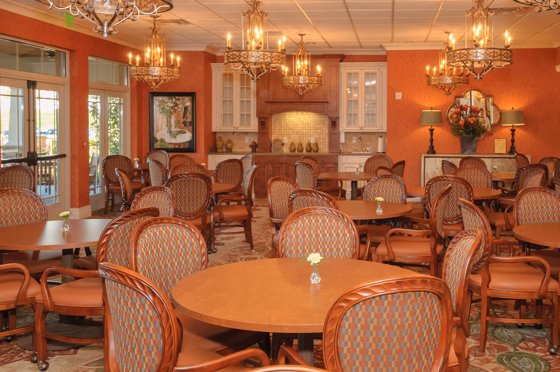 A dining room with multiple round wooden tables and patterned cushioned chairs. The room has warm orange walls, chandeliers hanging from the ceiling, and a kitchenette area with cabinets and a sink in the background. Small flower vases are placed on the tables.