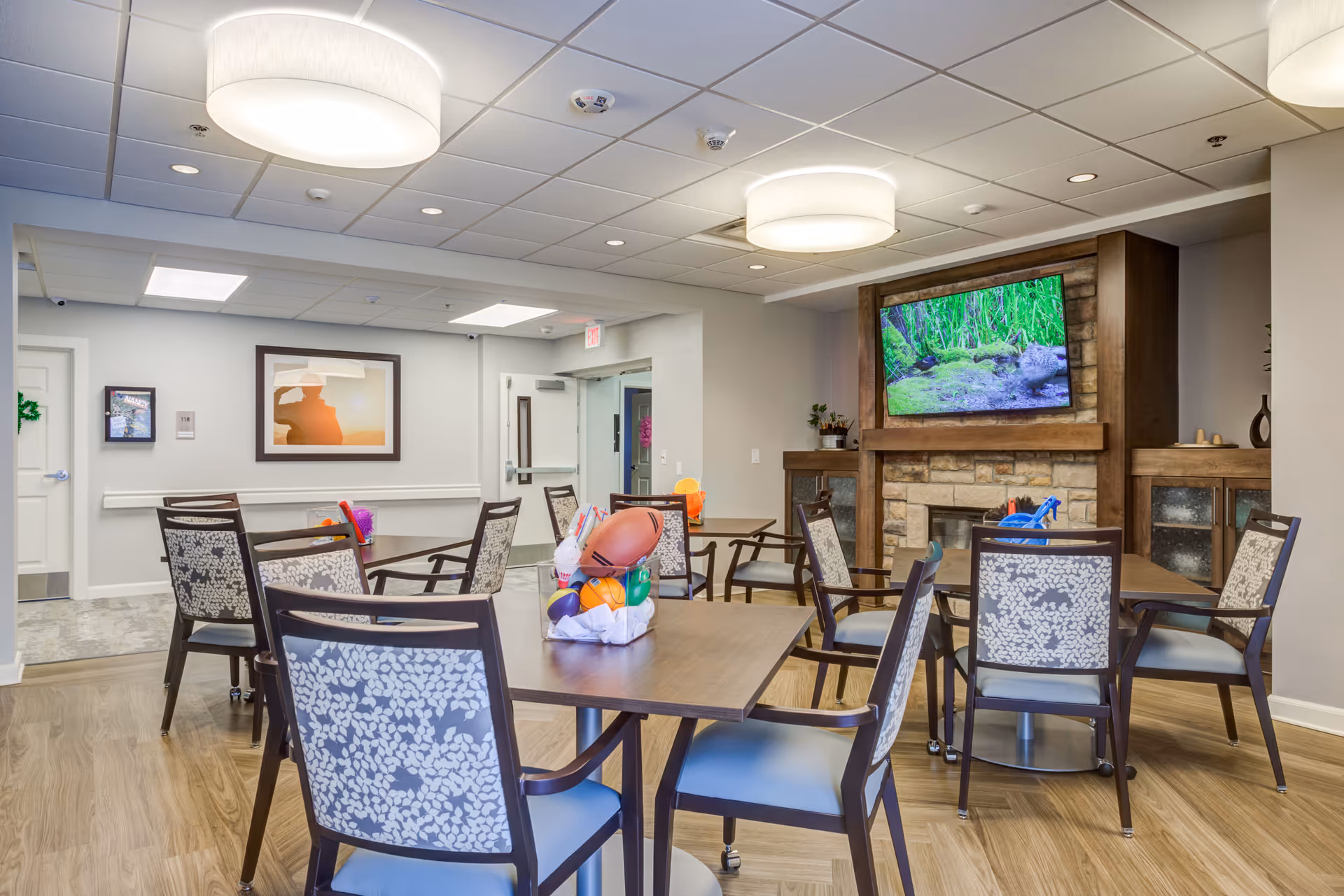Communal dining/activity room with tables and patterned chairs facing a wall-mounted TV above a stone fireplace.