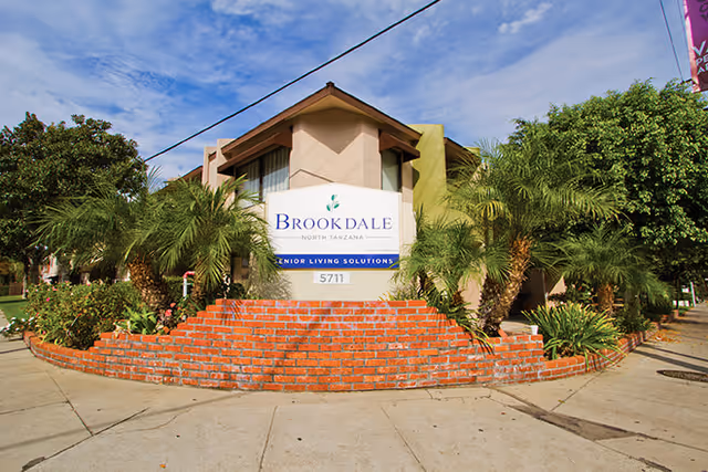 Exterior view of a senior living facility named Brookdale North Tarzana with a brick planter filled with palm trees and other greenery in front of the building under a partly cloudy sky.