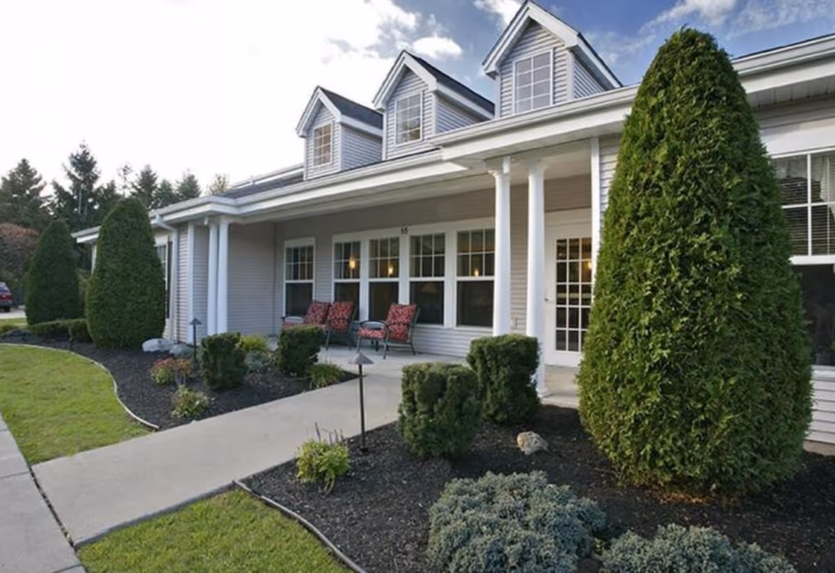 Exterior view of Tennyson Court facility showing a single-story building with gray siding, white columns, and three dormer windows. There is a concrete walkway leading to a porch with outdoor seating featuring red cushioned chairs. The landscaping includes neatly trimmed bushes, small trees, and a well-maintained lawn under a partly cloudy sky.