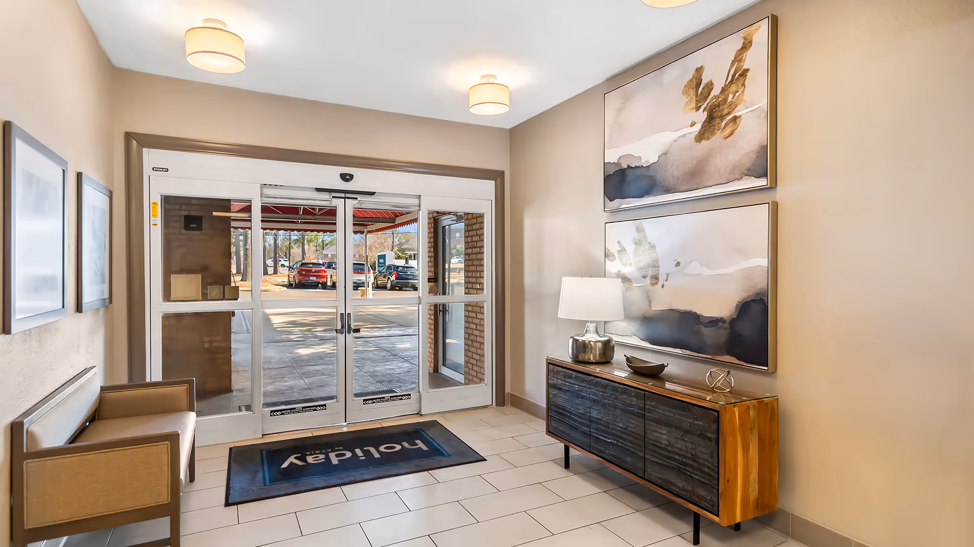 Entrance area of Holiday Chateau Ridgeland featuring glass automatic double doors with a view of parked cars outside. Inside, there is a bench on the left wall, two abstract paintings on the right wall above a wooden console table with a lamp and decorative items. The floor is tiled and the ceiling has two round light fixtures.