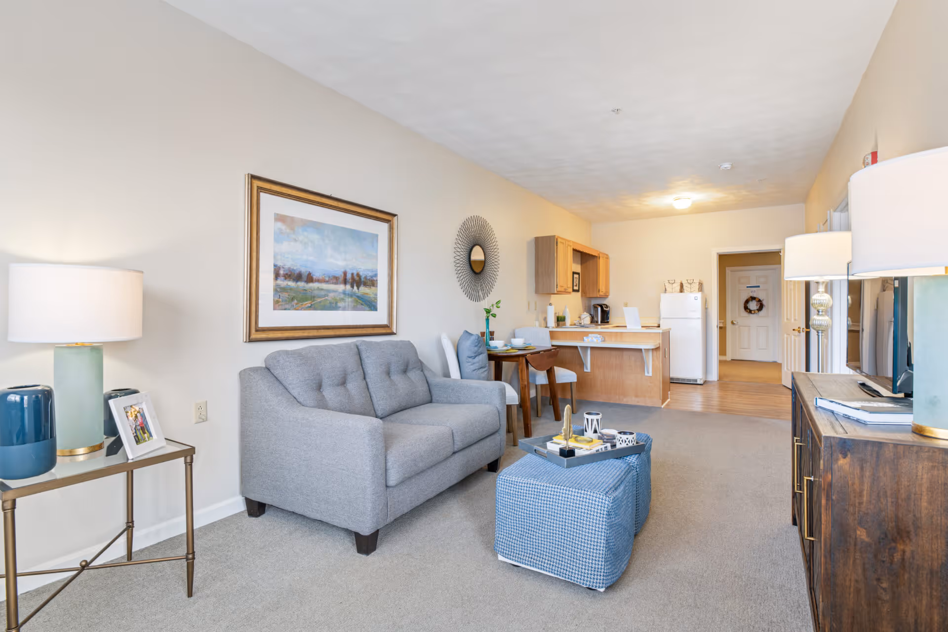 A bright and cozy living room area in a senior living facility with a gray sofa, a small dining table with two chairs, a blue ottoman with a tray on top, and a wooden cabinet with a TV. The room has beige walls, a framed landscape painting, a decorative round mirror, and two table lamps. The kitchen area with wooden cabinets, a white refrigerator, and a countertop is visible in the background.