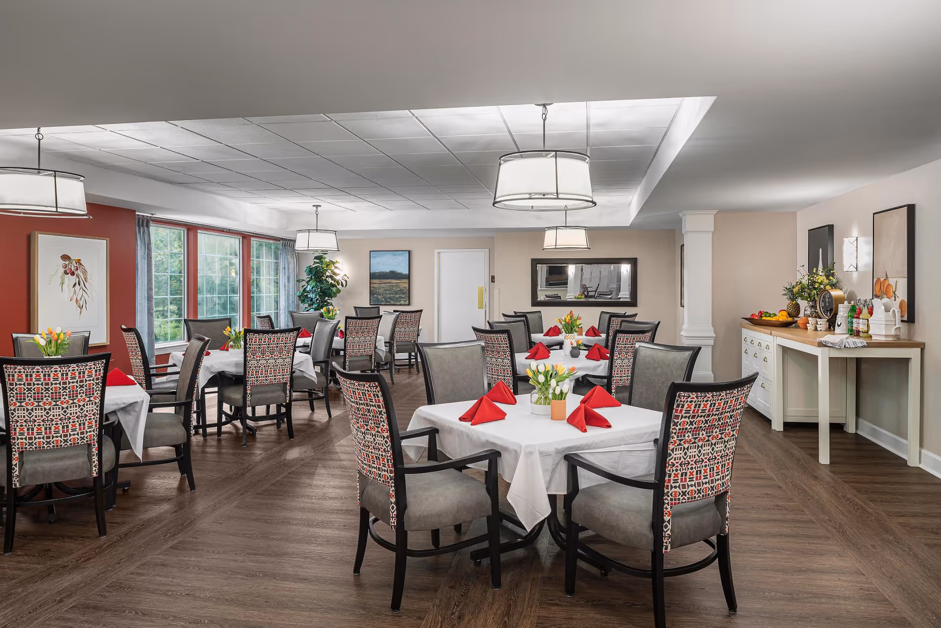 Bright dining room with multiple tables set with white tablecloths, red napkins, floral centerpieces, and patterned chairs.