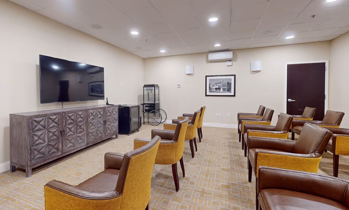 A senior living facility common room with two rows of brown and yellow armchairs facing a large flat-screen TV mounted on the wall above a decorative wooden cabinet. There is a popcorn machine and a small refrigerator against the far wall, along with a framed black and white picture and a closed dark wooden door.