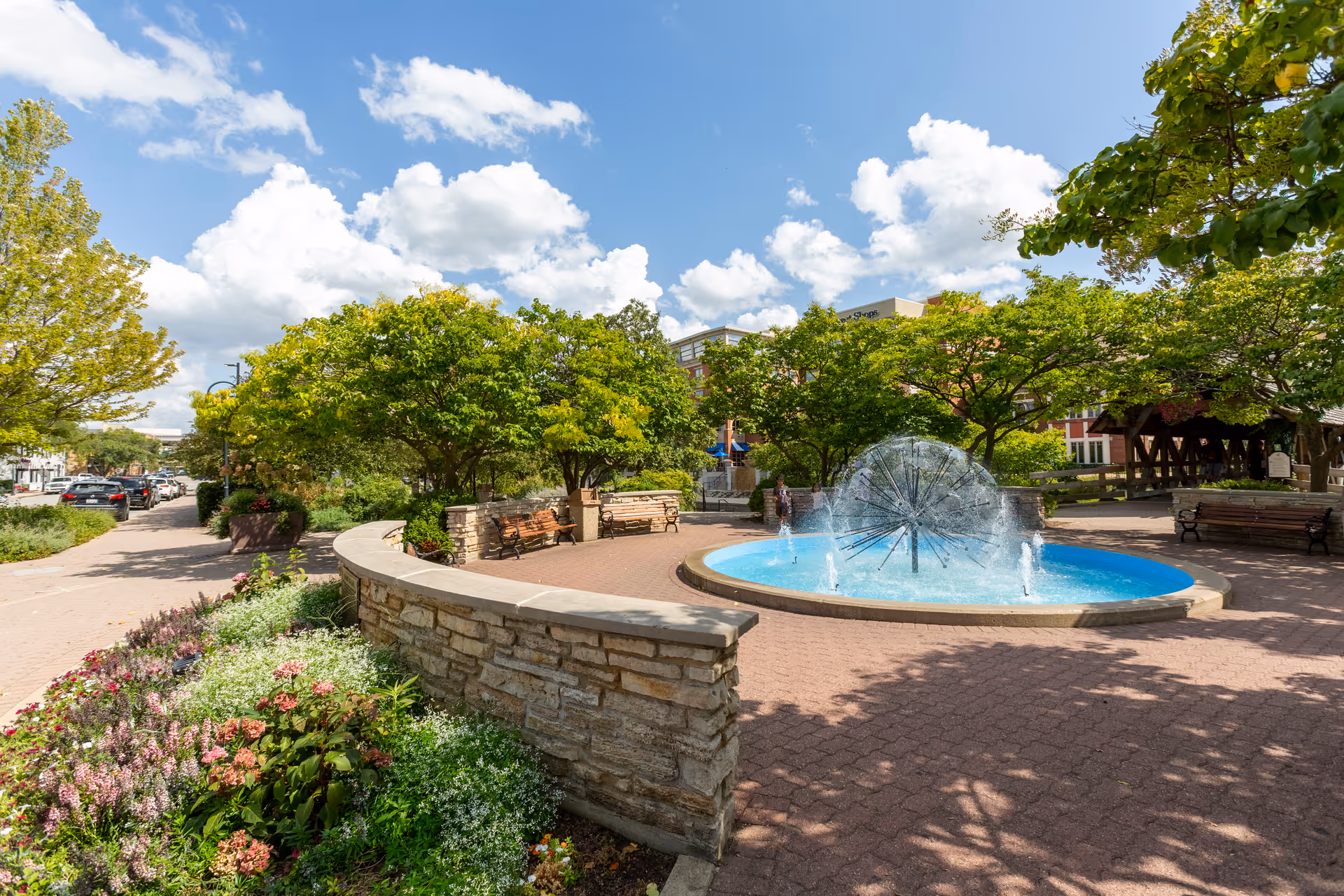 Outdoor garden area with a circular water fountain in the center, surrounded by trees, benches, and a stone wall. The sky is partly cloudy with blue patches visible.