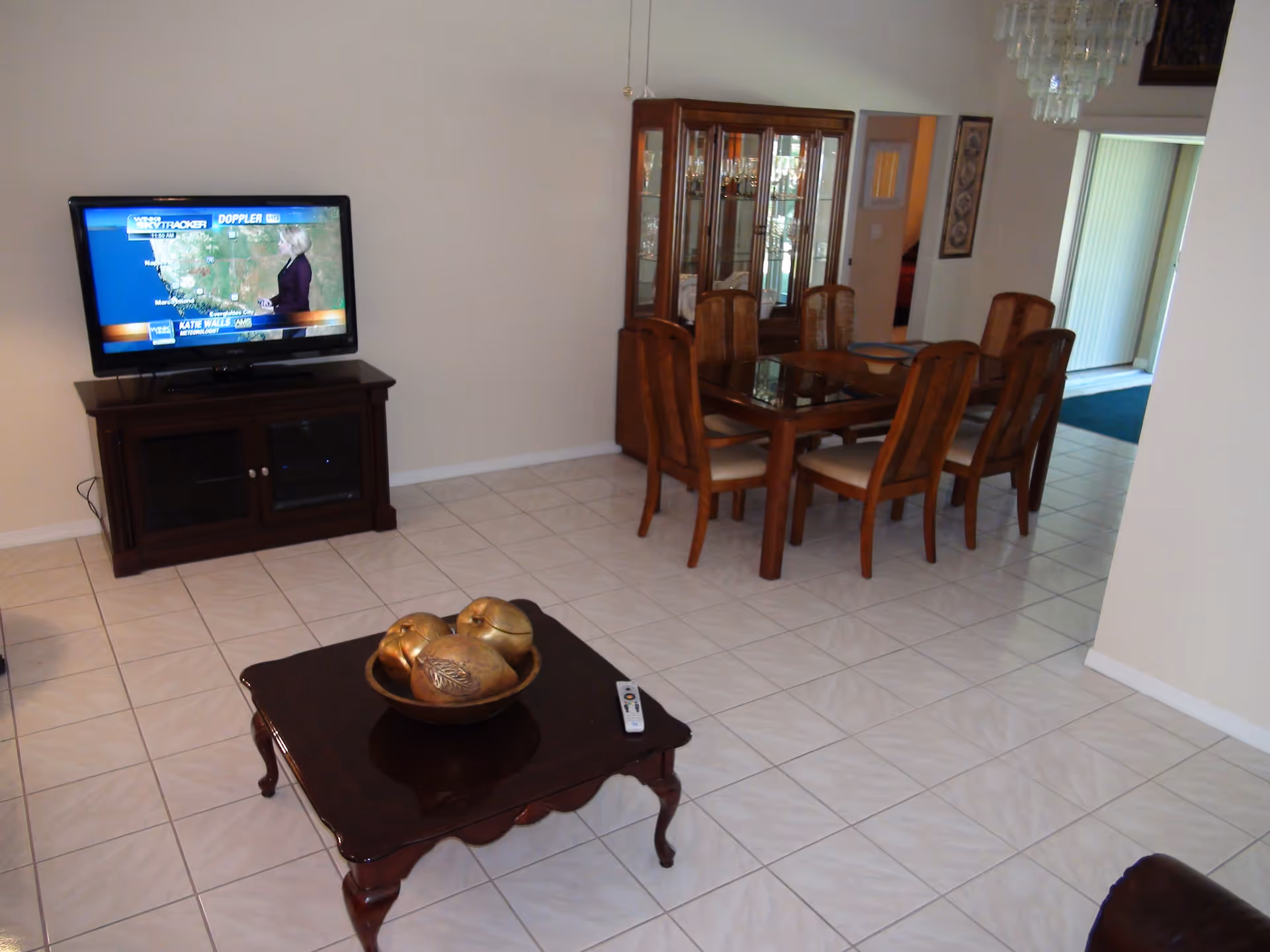 Interior view of a living and dining area with white tiled floors. A flat-screen TV on a dark wooden stand is showing a weather forecast. There is a wooden dining table with six chairs and a glass-fronted china cabinet behind it. A wooden coffee table with decorative golden apples and a remote control is in the foreground.
