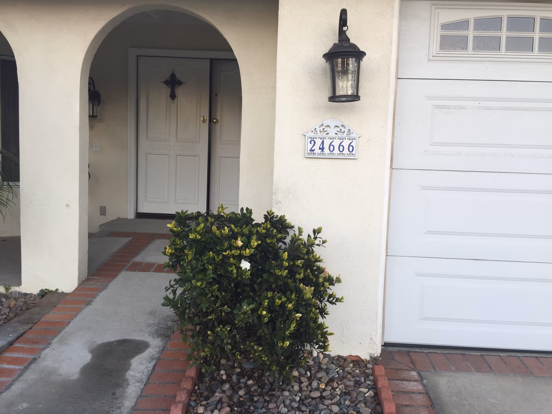 Front entrance of a house with a white double door featuring a decorative cross, an arched entryway, a wall-mounted lantern light, a house number plaque displaying 24666, a small green shrub with some white flowers, and part of a white garage door.