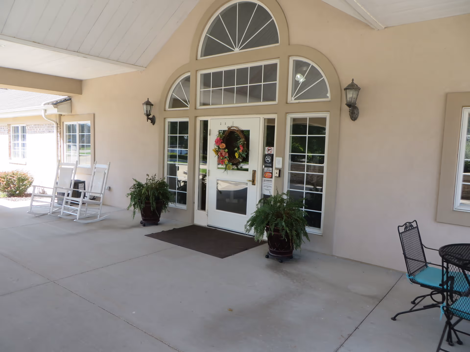 Covered entrance area of a building with a white door decorated with a floral wreath, flanked by tall windows and two potted plants. There are two white rocking chairs on the left side and a black metal table with chairs on the right side under the covered porch.