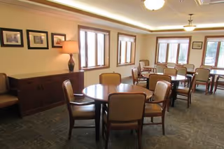 A well-lit dining room with multiple round tables surrounded by cushioned chairs. The room has several windows letting in natural light, framed pictures on the walls, a sideboard with a lamp, and a carpeted floor.