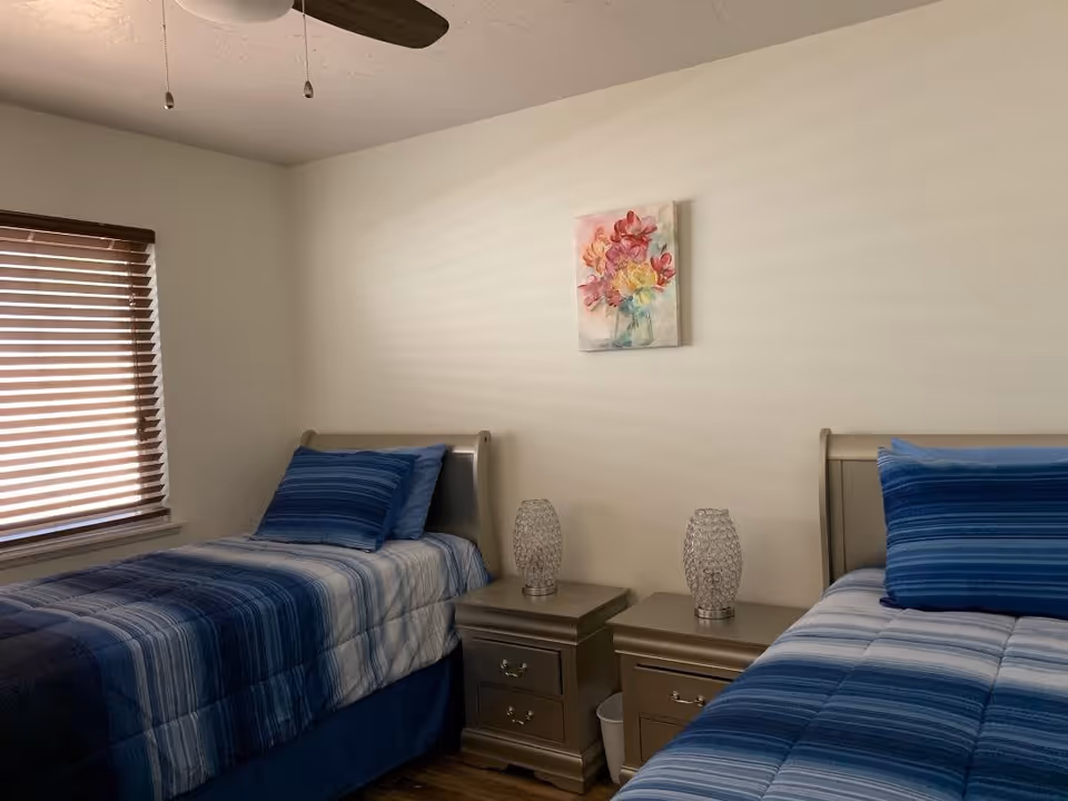 A bedroom with two twin beds, each with blue striped bedding and matching pillows. Between the beds are two nightstands, each with a decorative glass lamp. A window with wooden blinds is on the left wall, and a ceiling fan with pull chains is visible on the ceiling. A floral painting hangs on the wall above the nightstands.