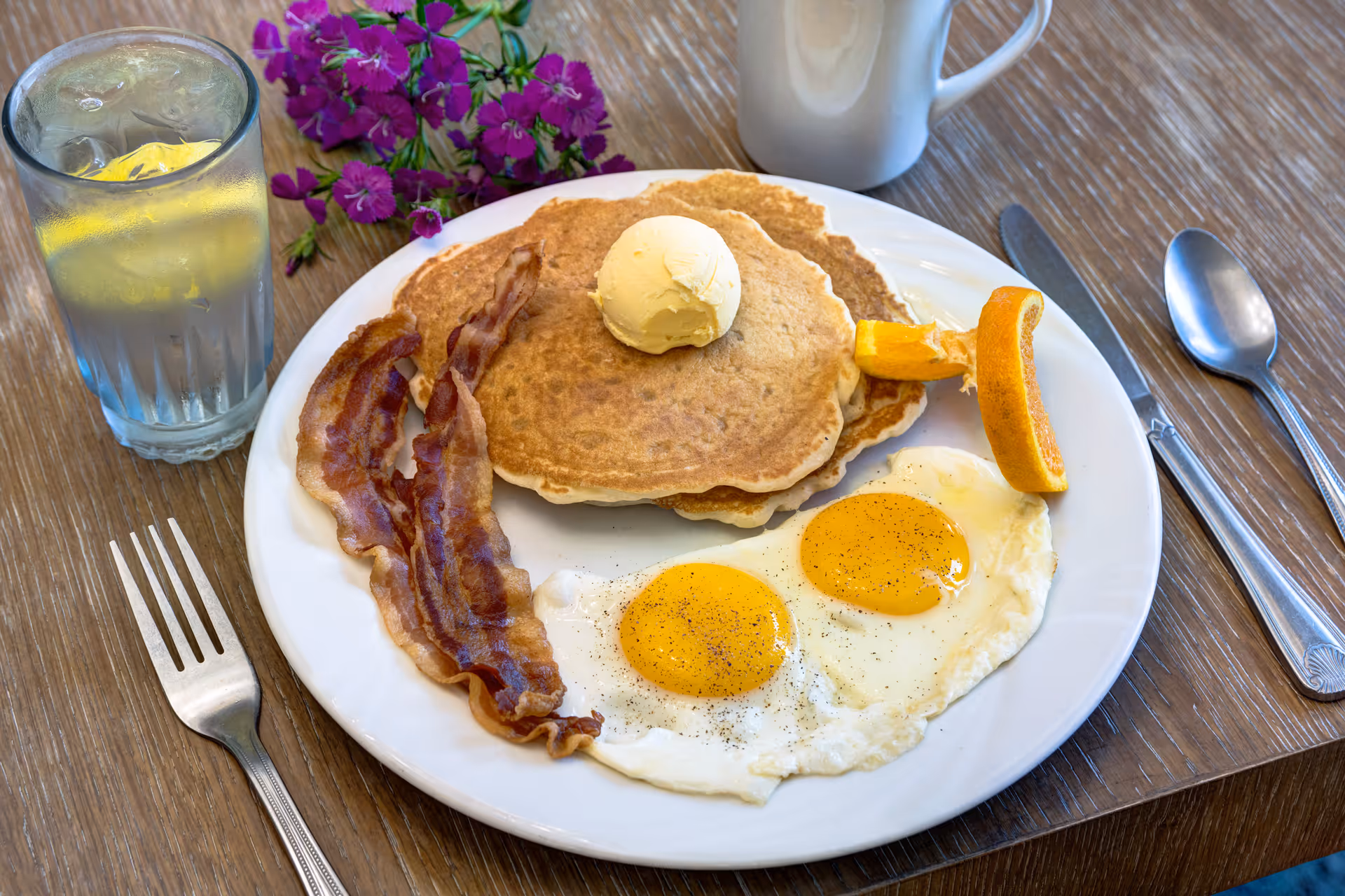 A breakfast plate with two sunny-side-up eggs sprinkled with black pepper, two strips of crispy bacon, two pancakes topped with a scoop of butter, and an orange slice. Next to the plate is a glass of water with a lemon slice and a white coffee mug on a wooden table with a purple flower in the background. Silverware including a fork, knife, and spoon are placed around the plate.