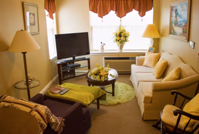 A cozy living room with a beige sofa adorned with yellow pillows, a dark armchair with a green throw blanket, a round glass coffee table with a floral centerpiece, a flat-screen TV on a wooden stand, two windows with red valances, a floor lamp, and a framed painting on the wall.