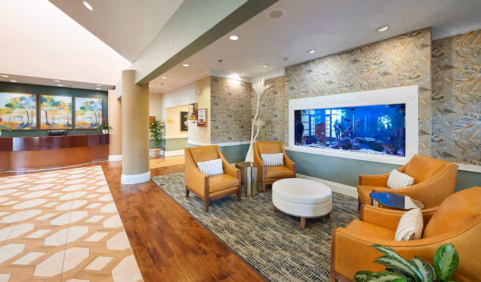 A bright and welcoming senior living facility lobby featuring a reception desk with three landscape paintings on the wall behind it. To the right, there is a cozy seating area with four tan leather armchairs, a round white ottoman, and a built-in aquarium set into a patterned wallpapered wall. The floor has a combination of wood and patterned tile, and there are plants and decorative elements throughout the space.