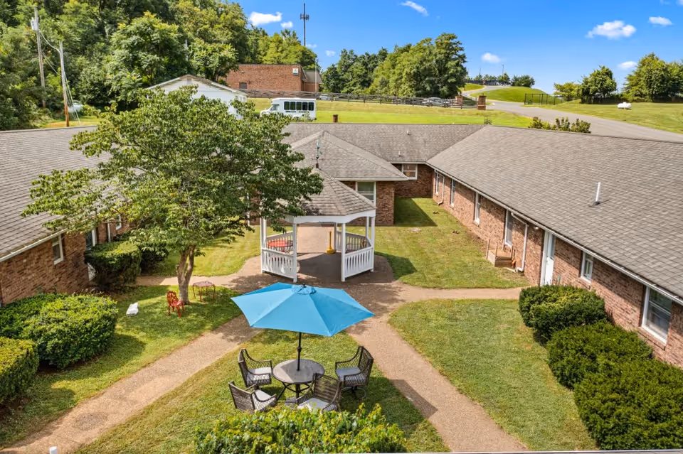 Outdoor courtyard area of Woods Cove Assisted Living featuring a gazebo, a tree, a table with four chairs under a blue umbrella, surrounded by brick buildings and green lawns under a clear blue sky.