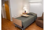 Single-occupancy bedroom with a hospital-style bed covered in a green blanket, a bedside table with a lamp and plant, and an upholstered recliner.