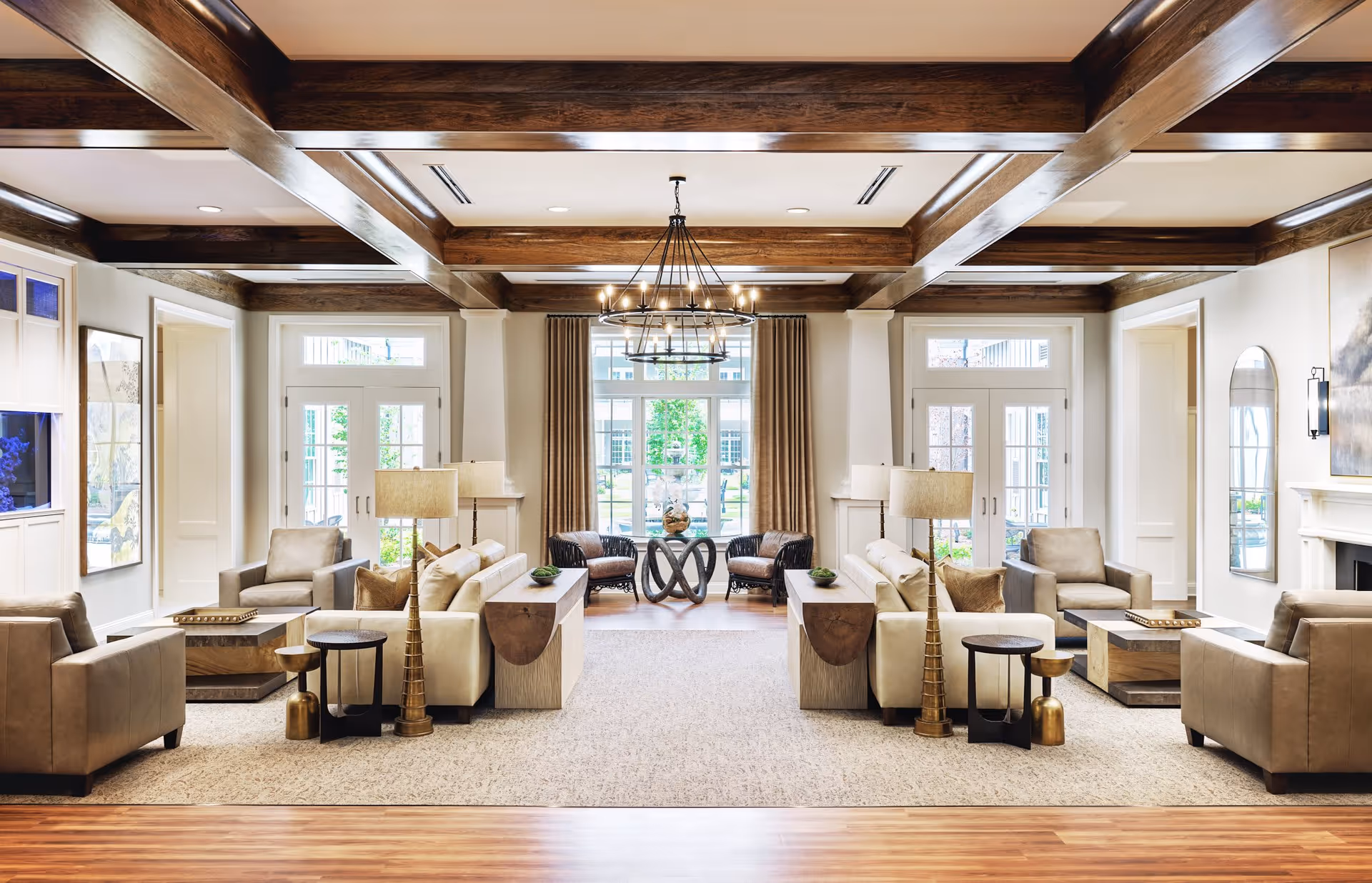 A spacious and elegant senior living facility common area with beige leather sofas and armchairs arranged around wooden coffee tables. The room features exposed dark wooden ceiling beams, a large chandelier, floor lamps, and large windows with beige curtains letting in natural light. There are decorative side tables and a view of an outdoor garden through the windows.