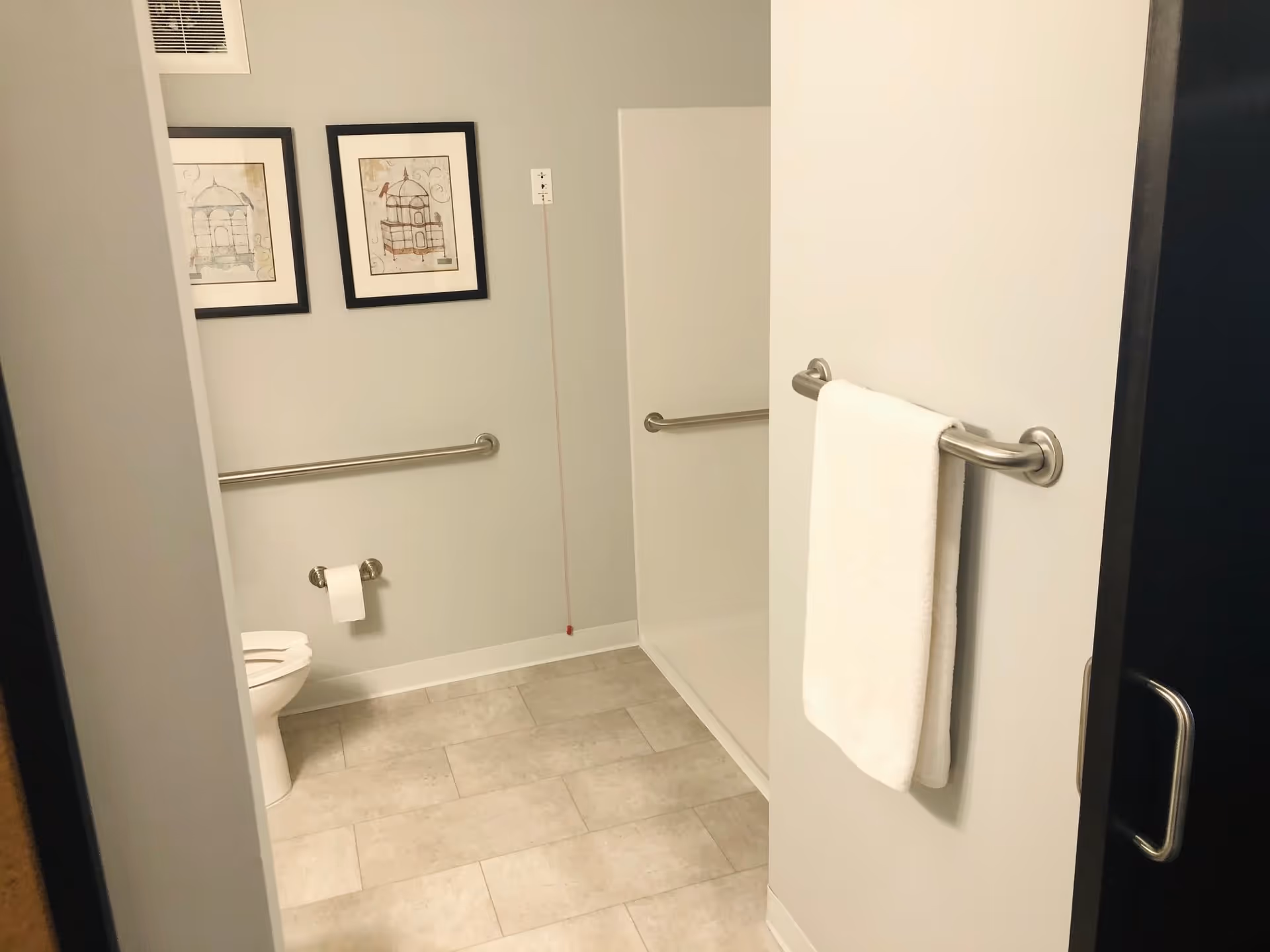 A clean and accessible bathroom with a toilet, grab bars on the walls, a white towel hanging on a bar attached to the door, and two framed pictures on the wall. The floor is tiled and the walls are painted light gray.