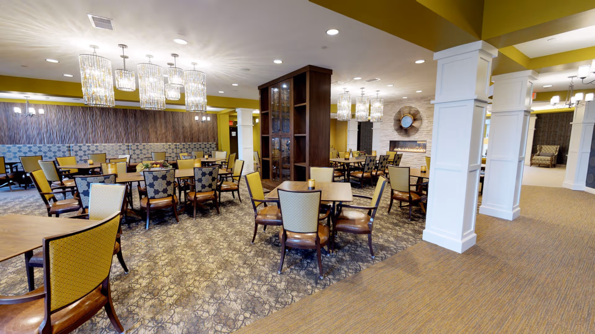 A spacious dining room in an assisted living facility with multiple wooden tables and chairs arranged neatly. The chairs have patterned upholstery in shades of yellow and blue. The room features modern chandeliers hanging from the ceiling, a decorative wooden partition in the center, and a fireplace with a circular mirror above it. The walls are painted in warm tones with white columns and patterned carpeting covering the floor.