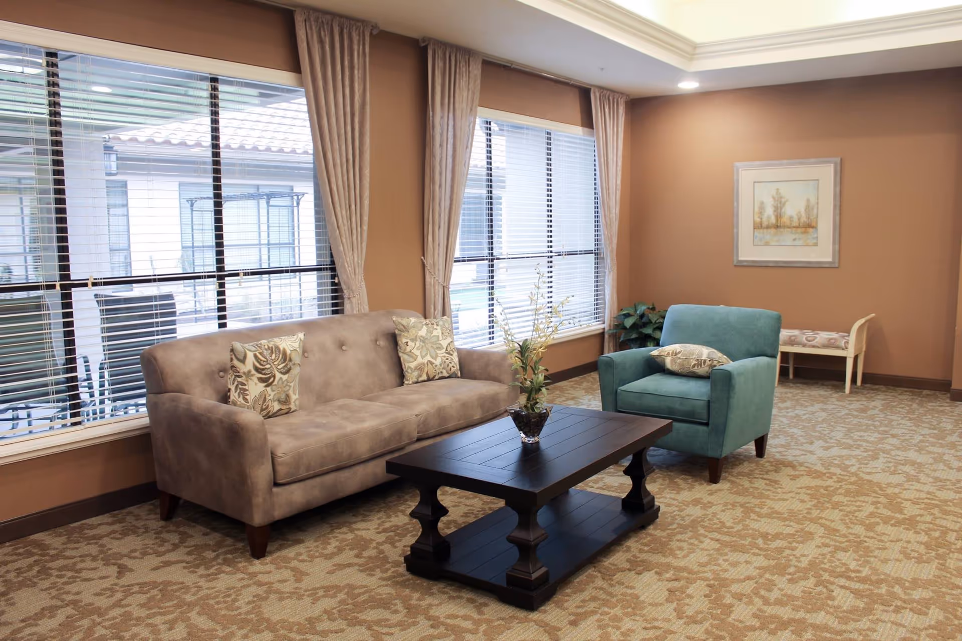 A cozy living room area with a beige sofa adorned with two patterned pillows, a teal armchair with a matching pillow, and a dark wooden coffee table with a small plant in a glass vase. The room has large windows with blinds and beige curtains, a patterned carpet, a framed artwork on the wall, and a small bench with a cushion in the corner.