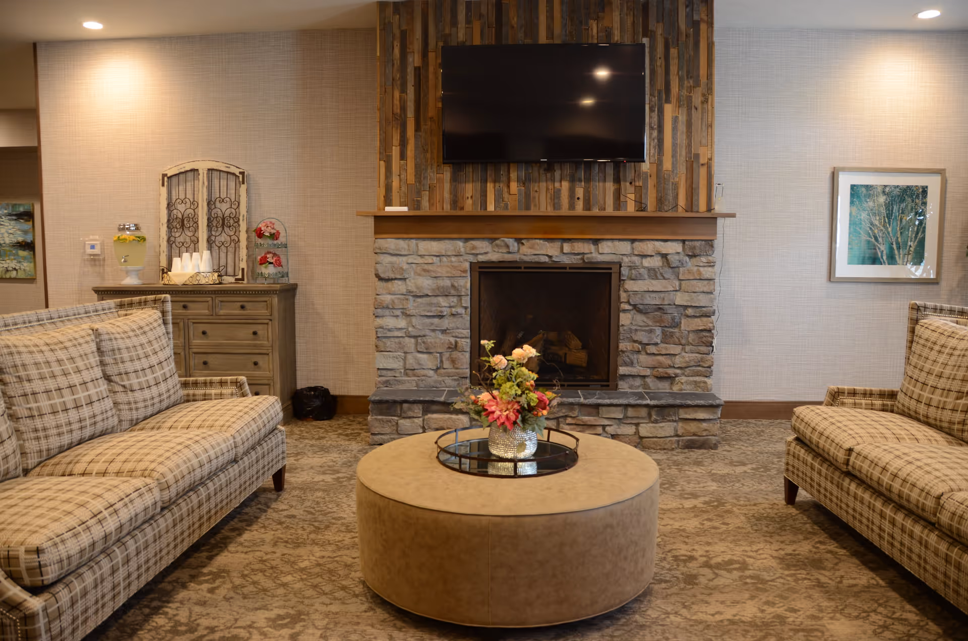 A cozy living room area with two plaid sofas facing each other, a round ottoman with a flower arrangement on top, a stone fireplace with a wooden mantel, and a flat-screen TV mounted above the fireplace. There is a wooden cabinet with a beverage dispenser and cups on the left side and a framed picture on the right wall.